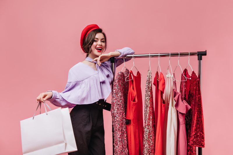 Dark-haired woman with red lipstick smiles, leans on stand with clothes and holds package on pink background